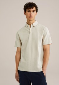 Polo de culoare crem cu mânecă scurtă, cu un model texturat, cu un guler clasic și o închidere cu trei butoane. Asortat cu pantaloni închiși la culoare.