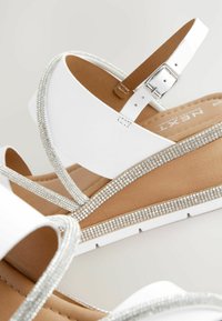 Sandales à talons compensés blanches en matériau lisse, ornées de strass scintillants le long des brides et de la semelle. Boucle au niveau de la cheville réglable.