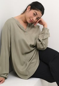 Olivově zelená halenka s dlouhými rukávy, véčkovým výstřihem a sklady na rukávech, v kombinaci s černými kalhotami. Hladká textilie s volným střihem.