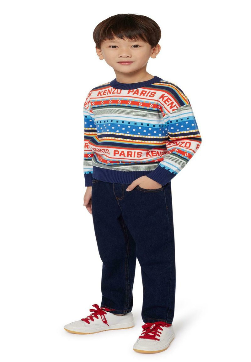 Pull en laine coloré avec des rayures bleues, rouges et à motifs, associé à un jean en denim foncé et des baskets blanches avec des lacets rouges.