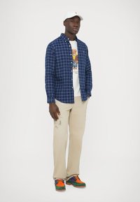 Marinblå rutig skjorta med knappar över en vit t-shirt med tryck, beige byxor och färgglada sneakers. En ljus hatt på huvudet.