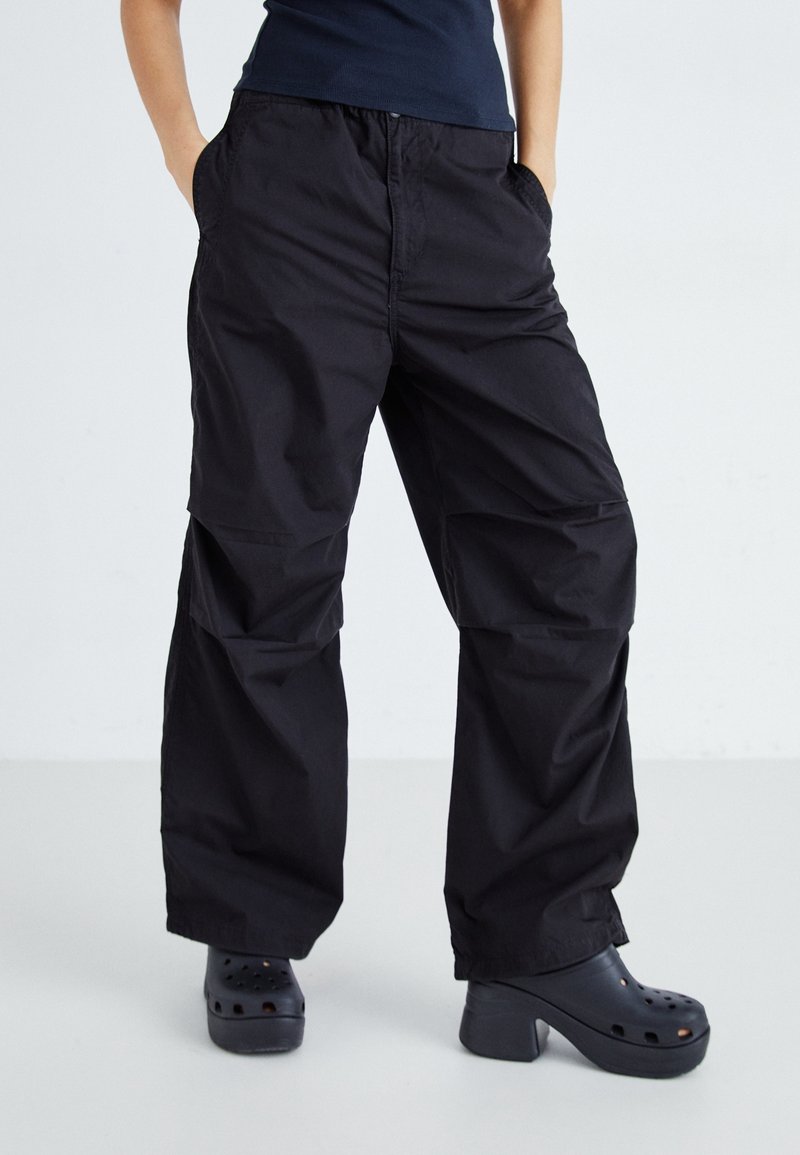 Personne portant un pantalon cargo noir ample avec des poches latérales et des sabots à plateforme noire épaisse, les mains dans les poches, debout devant un fond uni.