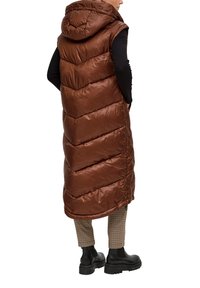 Person, der eine lange braune gepolsterte ärmellose Kapuzenweste, ein schwarzes langärmliges Oberteil, karierte Hosen und schwarze klobige Stiefel trägt, von hinten betrachtet.
