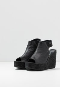 Sandalias cuña negras con punta abierta y tira trasera. Fabricadas en cuero texturizado con una base de yute trenzado y suela de goma.