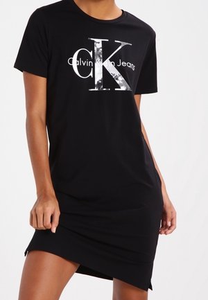 Frau trägt ein schwarzes Kurzarmkleid mit silbernem "Calvin Klein Jeans"-Logo auf der Brust und posiert mit den Händen in Hüfthöhe.