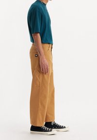 Tyrkysové tričko s krátkým rukávem v kombinaci s béžovými širokými kalhotami se bočními kapsami a nášivkou, doplněné černými teniskami.