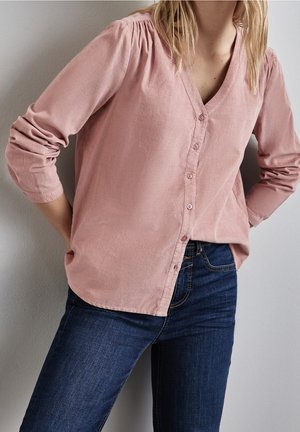 Kvinde iført en støvet lyserød langærmet skjorte med knapper og mørkeblå jeans, stående med hænderne på hofterne mod en lys væg.