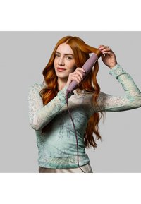 Hårstylingverktøy i en dempet lavendelfarge, med et teksturert grep og ventiler. Modellen har langt, bølgete rødt hår og har på seg en mønstret mintfarget skjorte.