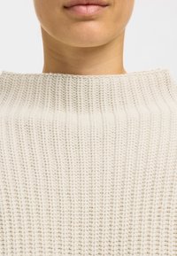 Pull tricoté couleur crème avec un col montant côtelé et une texture fine, montrant un gros plan sur l'encolure et la zone de la poitrine.