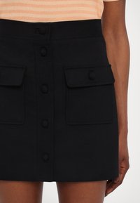 Zwarte minirok met een rechte snit, twee knoopzakken, knopen aan de voorkant en een soepele textuur. De stof lijkt gestructureerd en stevig te zijn.