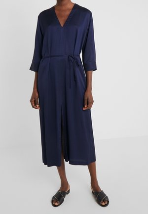 Mujer con vestido midi azul marino con escote en V, mangas tres cuartos, cinturón en la cintura, abertura frontal y sandalias negras sin cierre.