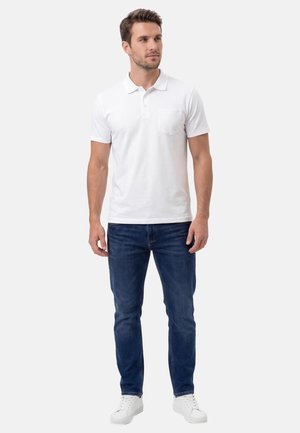 Mann steht und trägt ein weißes Polohemd mit kurzen Ärmeln, dunkelblaue Jeans und weiße Turnschuhe, blickt leicht zur Seite.