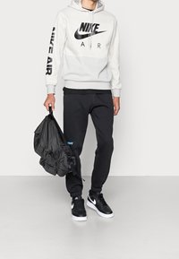 Světle šedá Nike Air mikina s černým logem, spárovaná s černými tepláky a černými teniskami. Jednotlivec drží černou tašku.