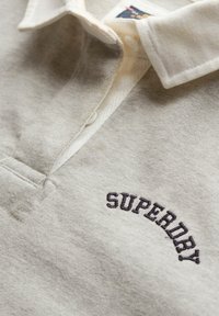Polo de color gris claro hecho de una tela suave. Presenta una tapeta abotonada y un logotipo bordado de "SUPERDRY" en azul oscuro. Diseño con cuello.