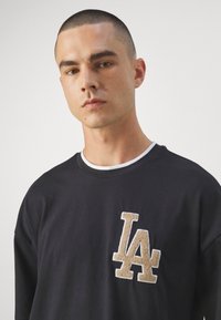 Sudadera negra con cuello interior blanco, con un logotipo difuso en beige "LA" en el lado izquierdo del pecho. Tejido suave, ajuste relajado.