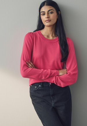 Maglione rosa brillante con scollatura rotonda e maniche lunghe, abbinato a jeans neri. Tessuto liscio, accenti a coste sottili sui polsini.