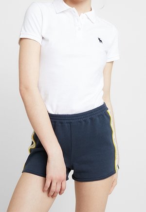 Biała koszulka polo z kołnierzykiem i małym logo, połączona z granatowymi szortami z żółtym paskiem z boku. Wygodne dopasowanie i gładka tekstura.