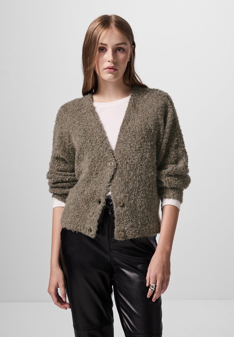 Luddig olivgrön cardigan med V-ringning, två knappar och lång ärm, som bärs med en vit topp och svarta läderbyxor.