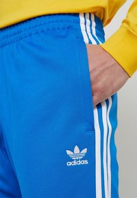Blå träningbyxor med vit adidas-logotyp, elastisk midja och sidofickor. Har tre vertikala vita ränder längs utsidan av benet.