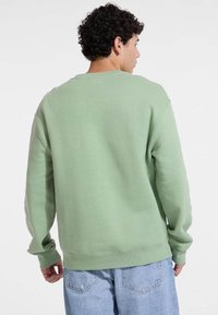 Zelena majica s okruglim izrezom, dugim rukavima, rebrastim manžetama i donjim rubom, napravljena od mekanog materijala, uparena s svijetloplavim trapericama.