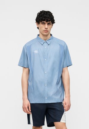 Jeune homme aux cheveux bouclés portant une chemise à manches courtes rayée bleue à boutons et un short noir avec des panneaux latéraux blancs.