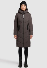 Vrouw in een lange, donkergbruine gewatteerde jas met capuchon, zwarte leggings, zwarte laarzen en een zwarte gebreide muts tegen een eenvoudige achtergrond.