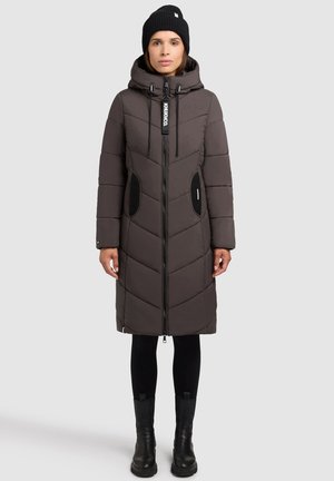 Vrouw in een lange, donkergbruine gewatteerde jas met capuchon, zwarte leggings, zwarte laarzen en een zwarte gebreide muts tegen een eenvoudige achtergrond.