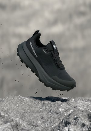 Pantof de alergare trail Mammut negru cu talpă groasă și aderență robustă, suspendat deasupra unui teren stâncos cu mici resturi care cad.