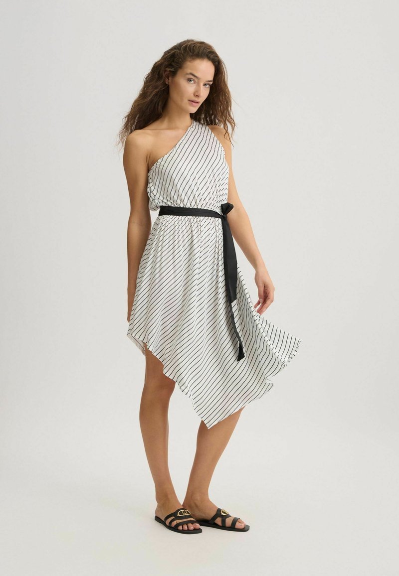 Weißes asymmetrisches Kleid mit schwarzen Streifen, ein Schulterdesign, tailliert mit einem schwarzen Gürtel und fließendem Saum. Getragen mit schwarzen Sandalen.