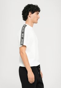 Jaunas vyras baltame marškinėlyje su juodu "Versace Jeans Couture" logotipo juostele ant peties, derinamas su juodomis kelnėmis, stovintis šonu baltoje fone.