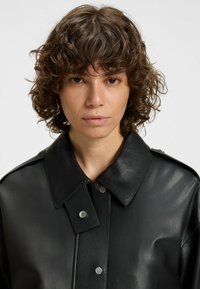 Black leather jacket featuring a large collar, snap-button closure, and smooth texture, with detailed stitching and metallic accents.