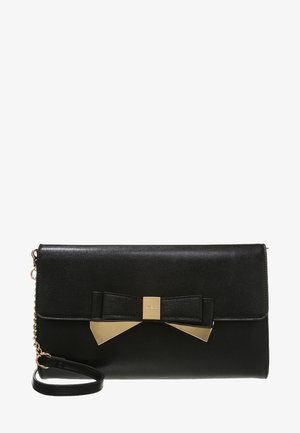 Bolso de mano de cuero negro con un detalle de lazo en tono dorado y una correa de muñeca desmontable, con cierre de solapa y un detalle de cadena en el lateral.