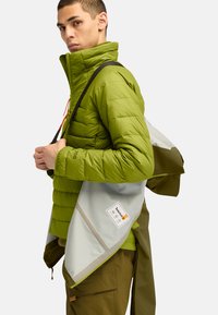 Chaqueta acolchada verde con cuello alto, que presenta un cierre frontal y una correa negra en el hombro. El forro interior es gris y cuenta con una etiqueta y detalles de bolsillo.