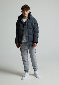 Abrigo acolchado gris oscuro con capucha, frente con cremallera y parche negro en el brazo, combinado con pantalones de chándal grises y zapatillas blancas. Diseño simple.