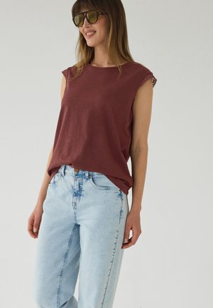 Kvinde iført brun ærmeløs top med blondekant og lyseblå højttaljede jeans, smilende og kigger til siden med solbriller på.