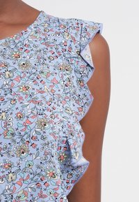 Blouse fleurie avec un fond bleu clair, présentant des manches volantées et un motif coloré de petites fleurs roses, jaunes et vertes.