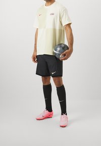 T-shirt color bloc crème clair et vert avec le logo Nike rouge, associé à un short noir, des crampons de football roses et des chaussettes noires jusqu'aux genoux, tenant un ballon gris.