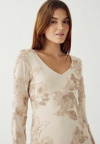 Fată tânără cu păr drept castaniu, îmbrăcată cu o rochie bej cu mânecă lungă și broderie florală texturată, privind înainte cu o expresie neutră.