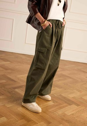 Persoon die een olijfgroene wijde cargobroek, witte sneakers, een wit shirt en een bruine leren jas met gouden armbanden draagt, staand op een houten vloer.