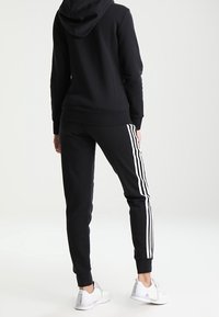 Haut à capuche noir avec capuche et pantalon de survêtement noir comportant trois rayures blanches sur les côtés, associés à des chaussures de sport blanches.