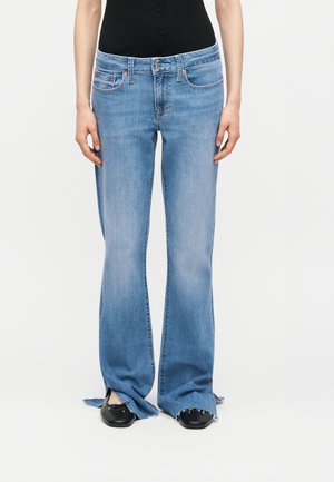 Frau trägt hellblaue ausgestellte Jeans mit ausgefransten Säumen, schwarze spitze Schuhe und ein eng anliegendes schwarzes Oberteil, steht vor einem schlichten weißen Hintergrund.