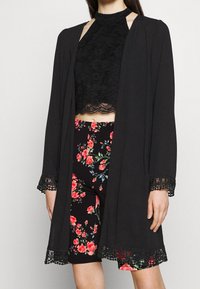 Svart kimono med spetsdetaljer över en svart blommig crop top och högmidjade shorts, med ett djärvt blommönster på mörk bakgrund.