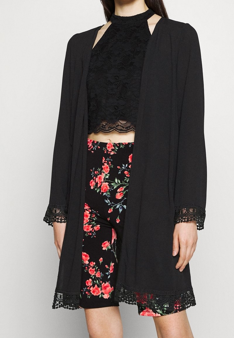Svart kimono med spetsdetaljer över en svart blommig crop top och högmidjade shorts, med ett djärvt blommönster på mörk bakgrund.