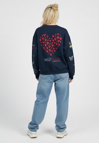 Una donna bionda in piedi di spalle, indossando un felpa blu navy con cuori rossi che formano un grande cuore e il testo "SWEET heart", jeans azzurri chiari e scarpe da ginnastica bianche.