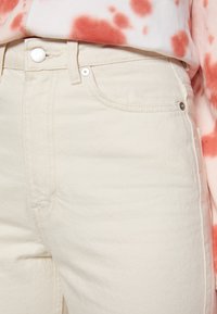 Jean en denim crème avec une coupe classique, présentant une poche avant, une fermeture à bouton en métal et des détails de couture discrets.