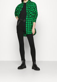 Camisa de cuadros verde y negra con dos bolsillos en el pecho, combinada con leggings negros y botas negras gruesas. De botones en el frente, con mangas largas.