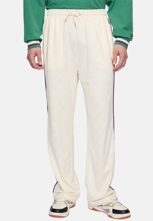 Cream-colored track pants with drawstring waist and purple side stripes worn with white and black sneakers, shown from waist to feet.