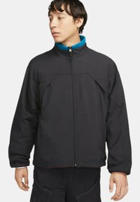 Svart isolerad jacka med hög krage och dragkedja. Har två framfickor och textilstruktur. Accentuerande blå foder är synligt.