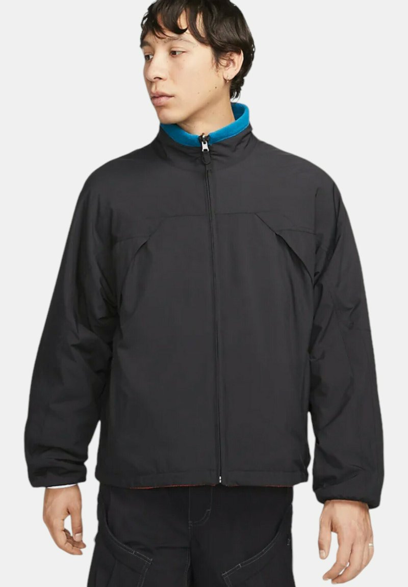 Svart isolerad jacka med hög krage och dragkedja. Har två framfickor och textilstruktur. Accentuerande blå foder är synligt.