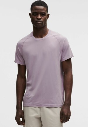 Mann trägt ein hellviolettes T-Shirt mit kurzen Ärmeln und rundem Ausschnitt sowie hellbeige Hosen, steht vor einem schlichten weißen Hintergrund.